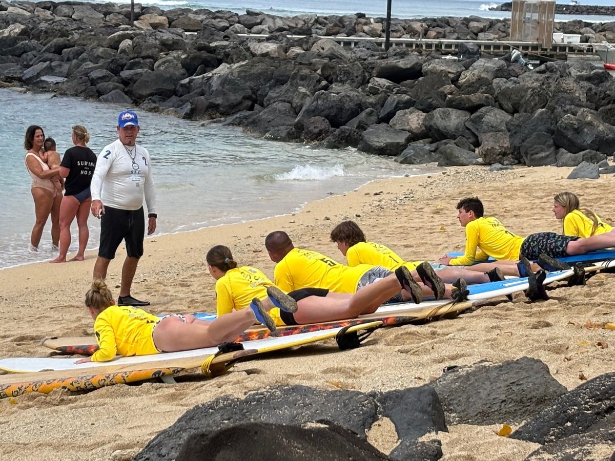 SFC students in yellow rash guards practicing paddle position on boards on the Waikīkī sand
